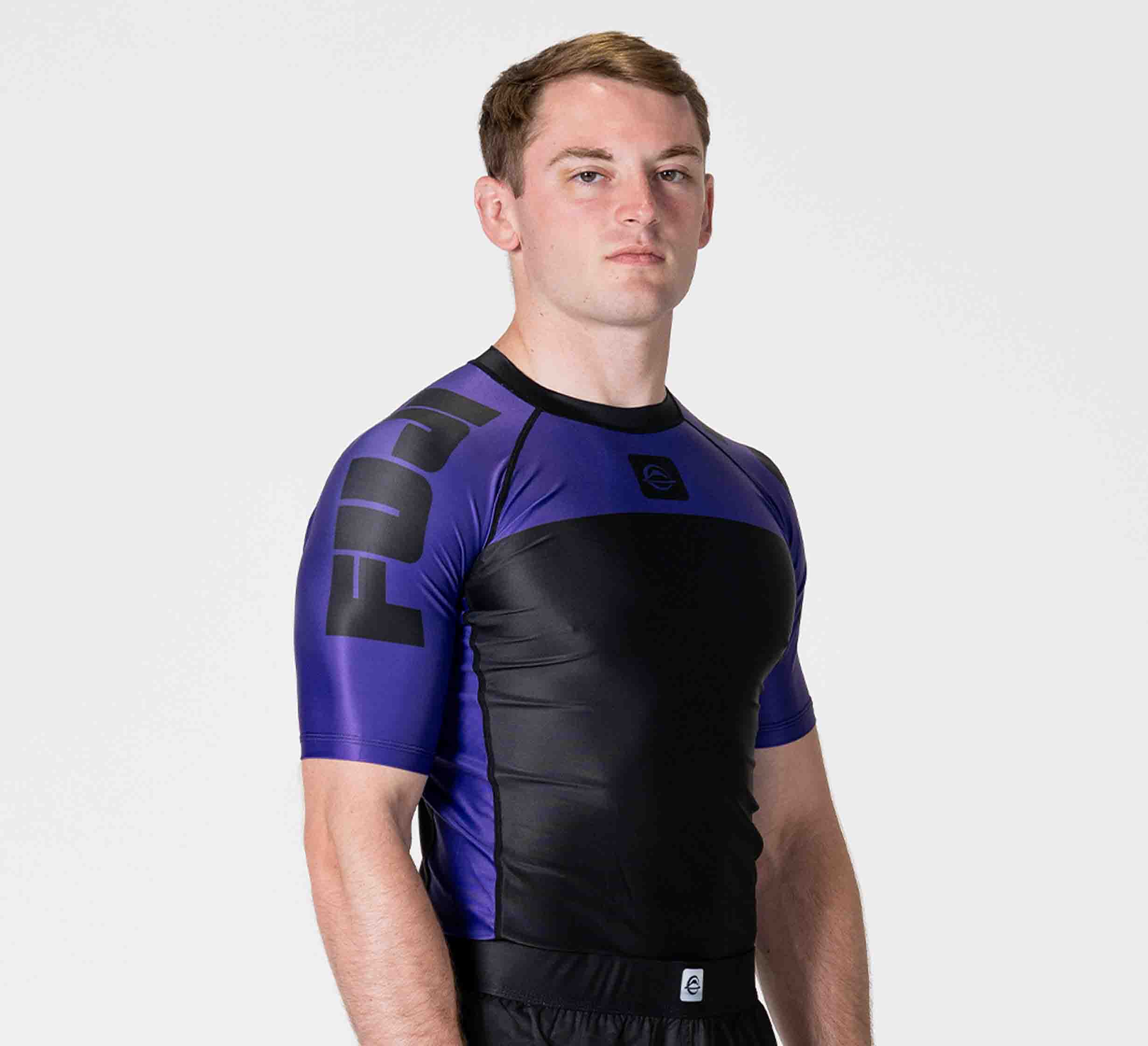 A man stands against a plain white background, wearing the Competition Ranked Rashguard Purple with FUJI on the sleeve. He lo