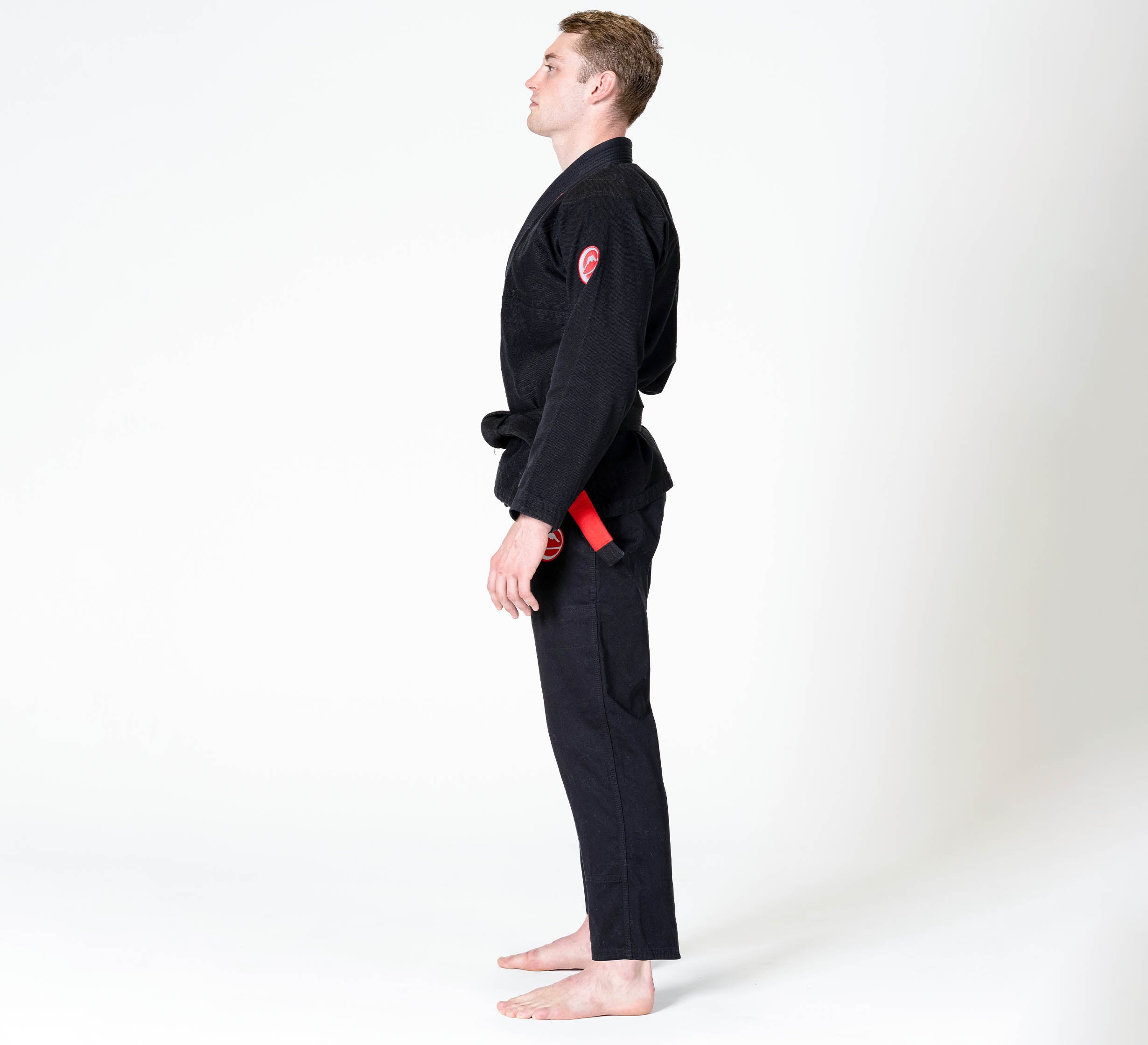 A person wearing the Competition BJJ Gi Black with a red and black belt stands barefoot in a side profile against a plain whi