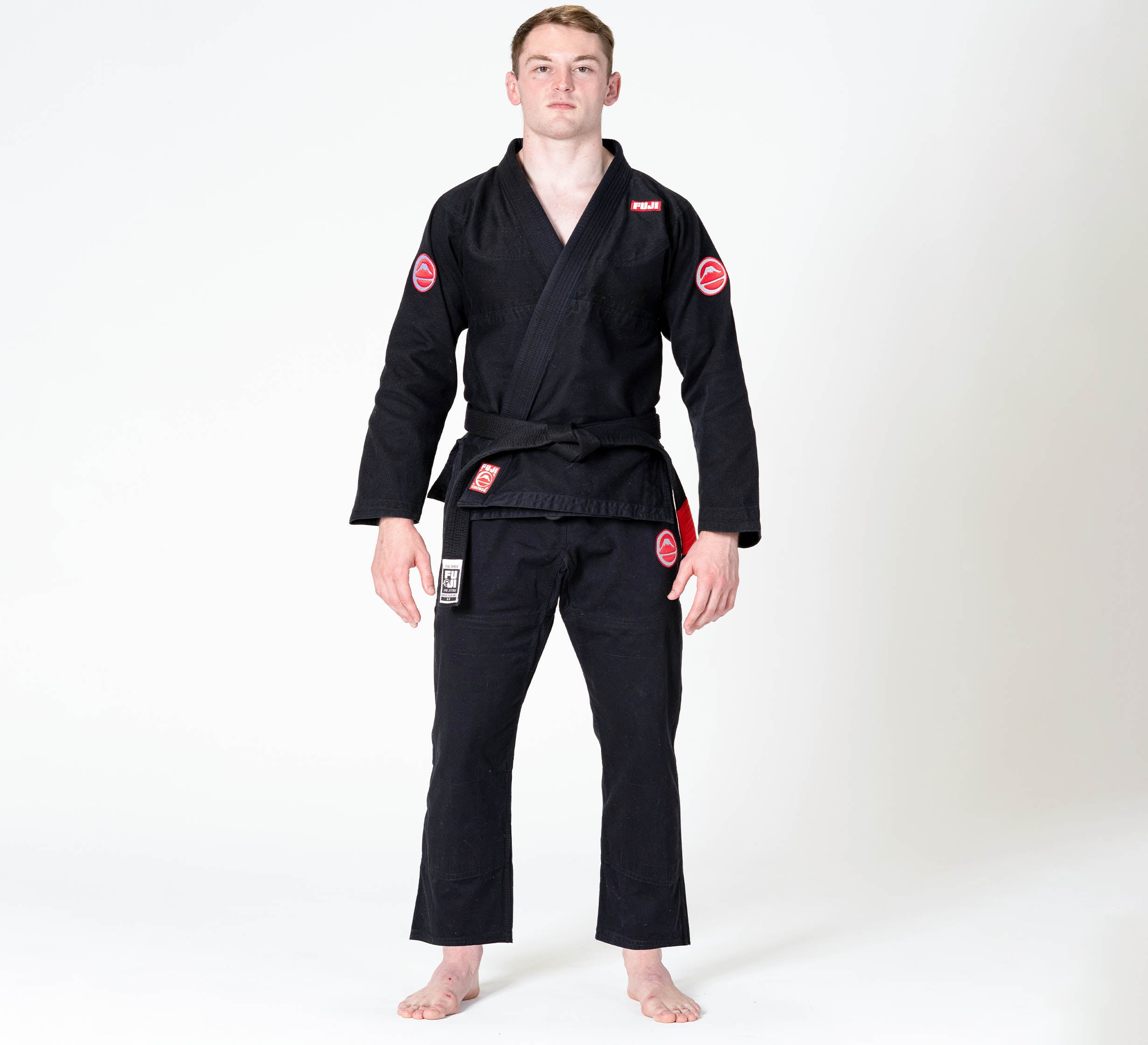 A person stands barefoot against a white background, wearing the Competition BJJ Gi Black with red and white patches and a bl