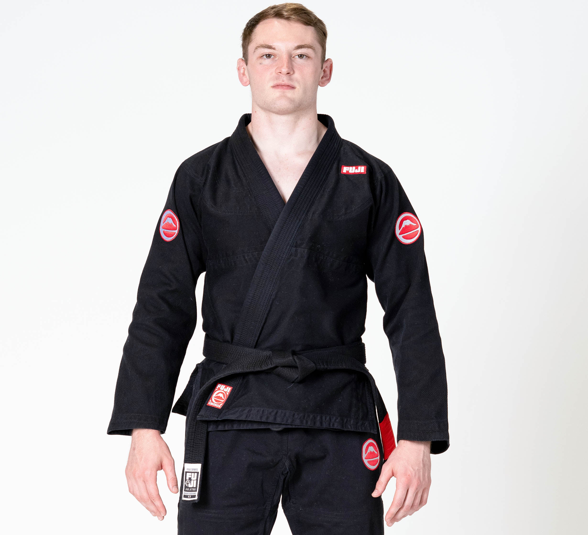 A person stands facing forward, wearing the Competition BJJ Gi Black with red patches and a black belt, set against a plain w