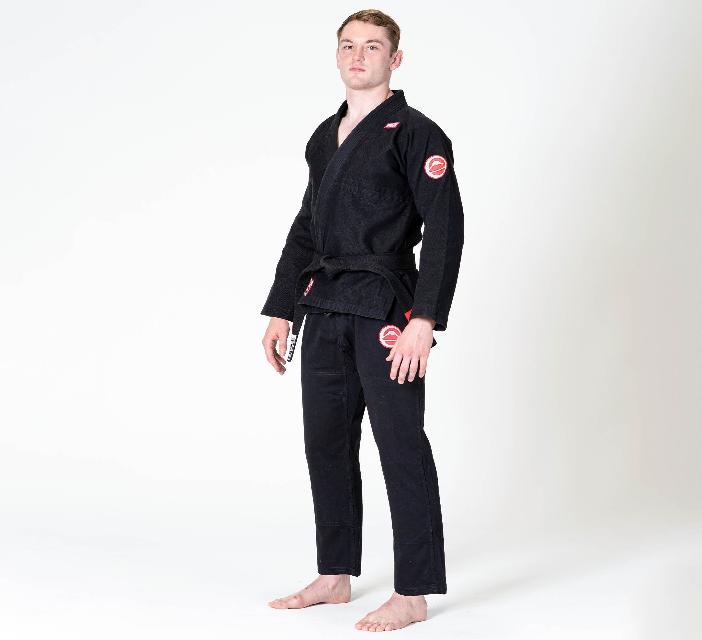 A person stands barefoot on a white background, wearing the Competition BJJ Gi Black with red patches and a black belt. They