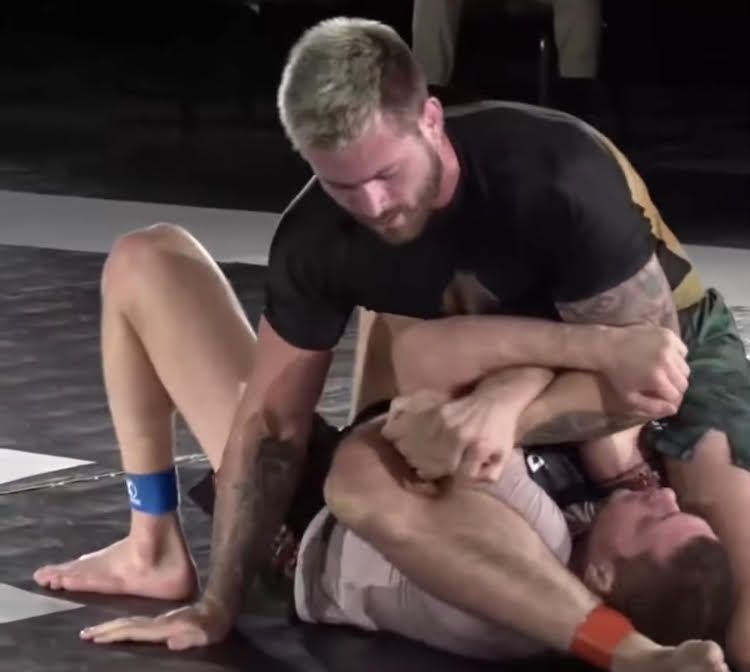 No-gi grappler in a black rashguard controlling an opponent, sweat-soaked training gear