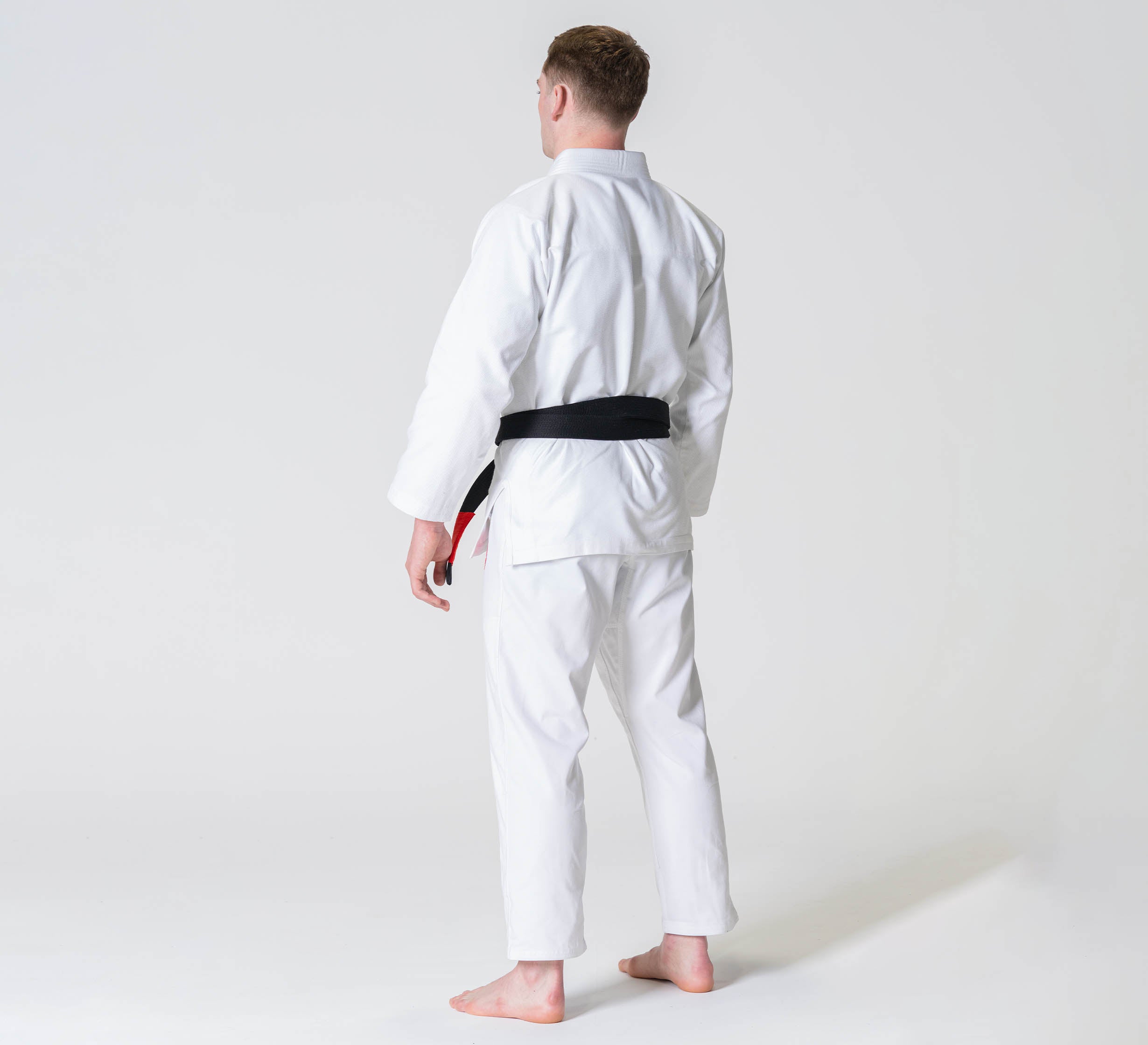 A person wearing the Competition BJJ Gi White with a black belt stands barefoot, facing away from the camera against a plain,