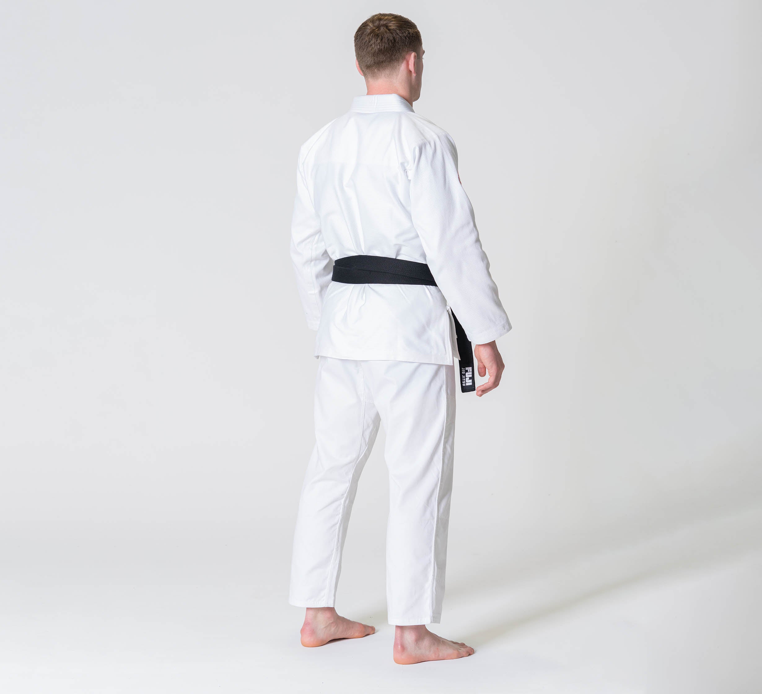 A person wearing the Competition BJJ Gi White, barefoot with a black belt, stands facing away from the camera against a plain