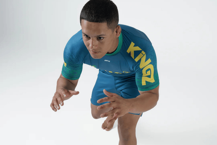 Athlete wearing a blue KINGZ rashguard and shorts in a low grappling stance on a white background.