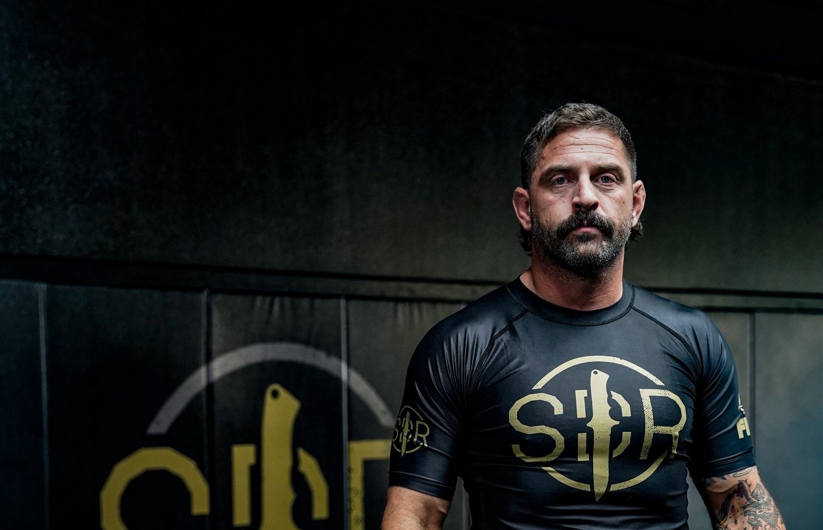 Grappler wearing a fitted black and gold competition rashguard in the gym, shoulder seams and sleeve construction visible