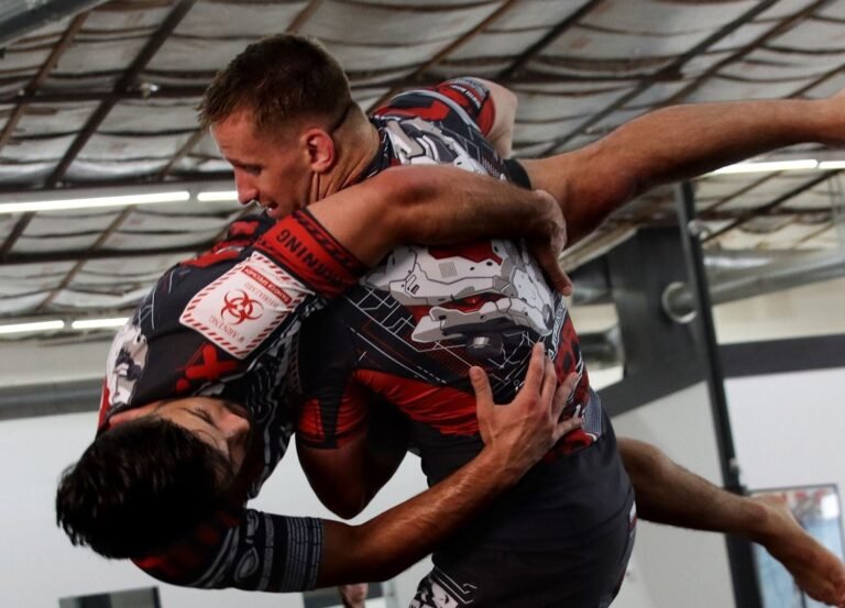 Two no-gi grapplers locked in a grip exchange, the heavily sublimated print on their rashguards visible exactly where hands are gripping the fabric