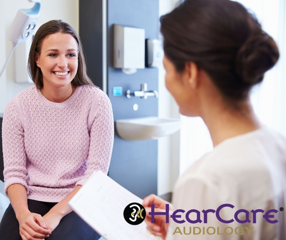 Smiling woman sits in clinic exam room as clinician holds paperwork and HearCare Audiology logo at b